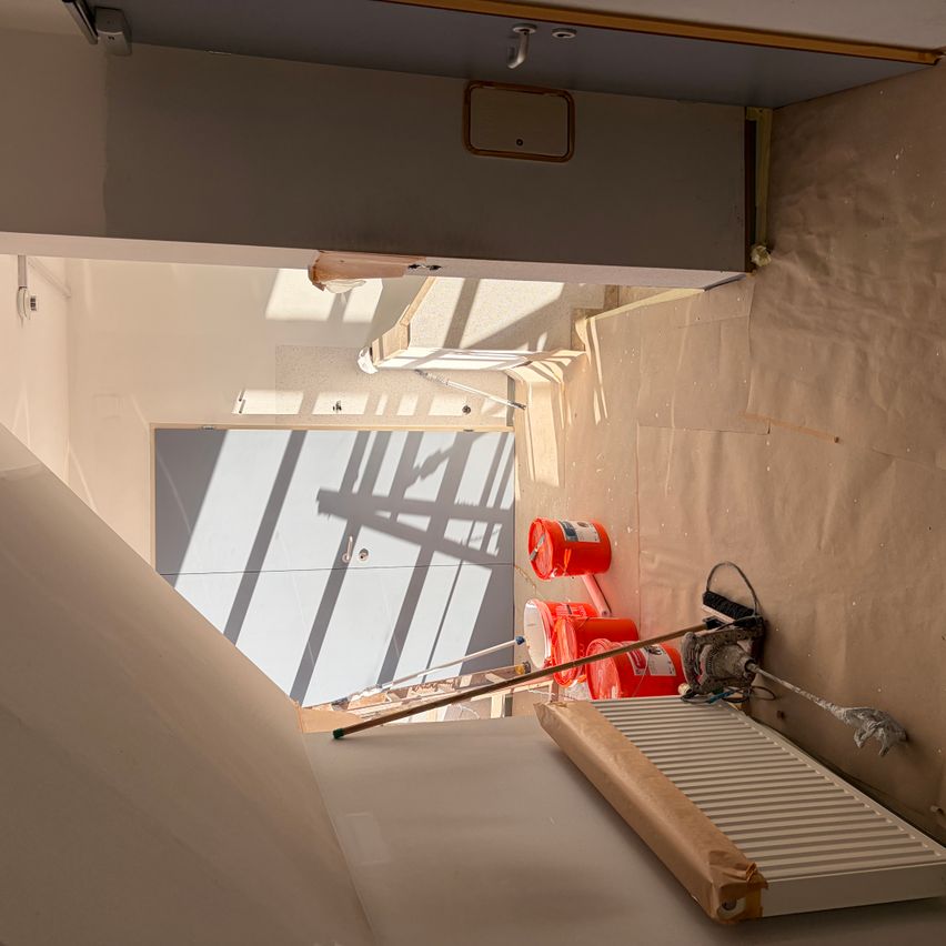 Ein Baustelle mit einer weißen Wand und einer Tür. Zwei orangefarbene Eimer stehen auf dem Boden. Ein weißer Heizkörper befindet sich auf der rechten Seite.
