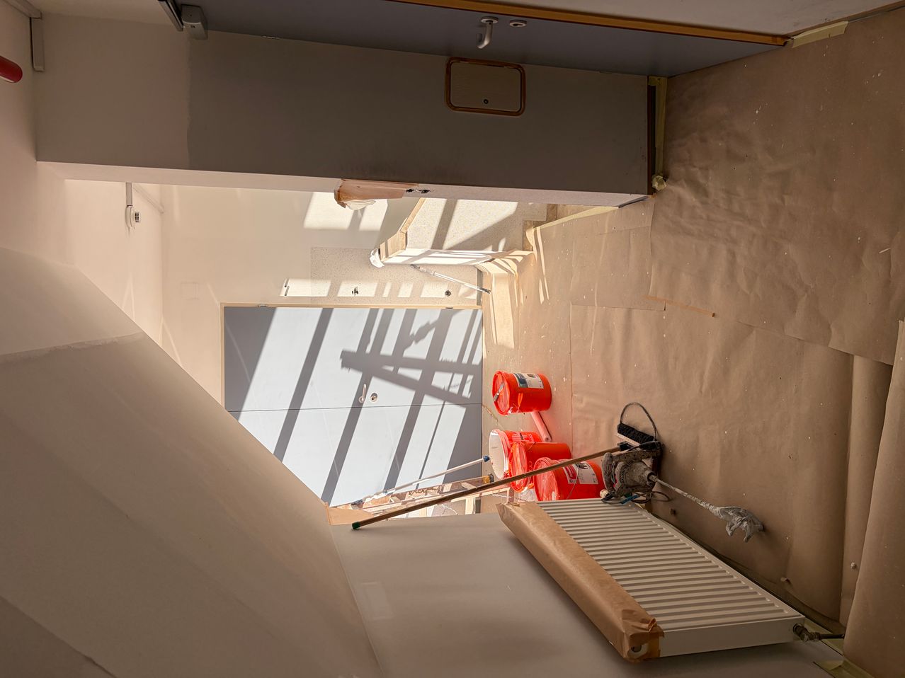 Ein Baustelle mit einer weißen Wand und einer Tür. Zwei orangefarbene Eimer stehen auf dem Boden. Ein weißer Heizkörper befindet sich auf der rechten Seite.