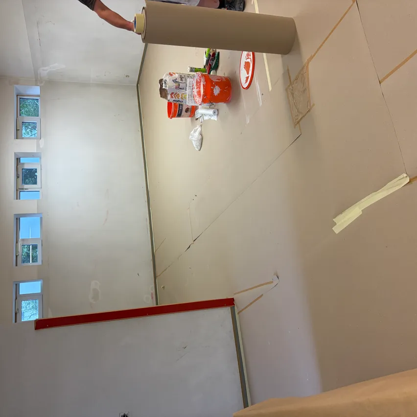 Ein Innenraum einer im Bau befindlichen Raum mit weißen Wänden, einem roten Rand und einer Person, die eine Rolle Klebeband hält. Es gibt zwei orangefarbene Farbbehälter, einen roten Teller und andere Materialien auf dem Boden.