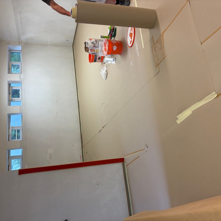 Ein Innenraum einer im Bau befindlichen Raum mit weißen Wänden, einem roten Rand und einer Person, die eine Rolle Klebeband hält. Es gibt zwei orangefarbene Farbbehälter, einen roten Teller und andere Materialien auf dem Boden.