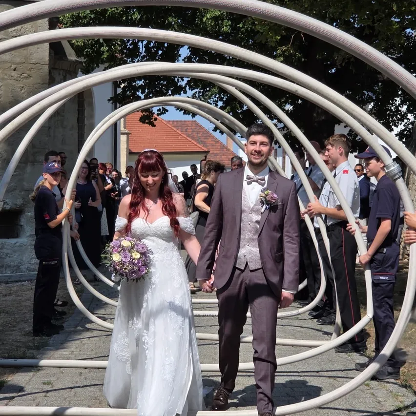 Ein frisch verheiratetes Paar geht durch eine einzigartige Bogenstruktur, umgeben von Zuschauern bei einer Hochzeit.