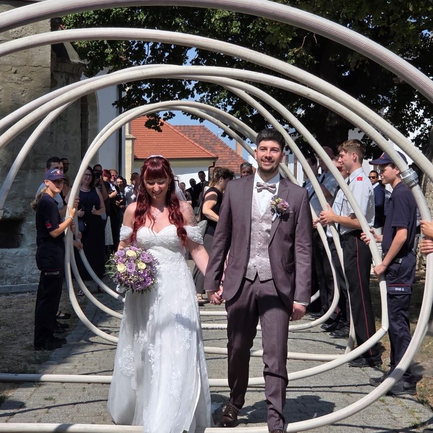 Ein frisch verheiratetes Paar geht durch eine einzigartige Bogenstruktur, umgeben von Zuschauern bei einer Hochzeit.