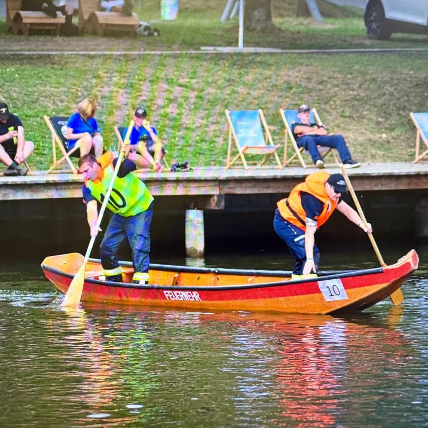 Zwei Männer in Warnwesten rudern mit einem roten und orangefarbenen Boot, das mit der Nummer 10 beschriftet ist, auf einem See, während Zuschauer auf Stühlen auf einem Steg im Hintergrund sitzen.