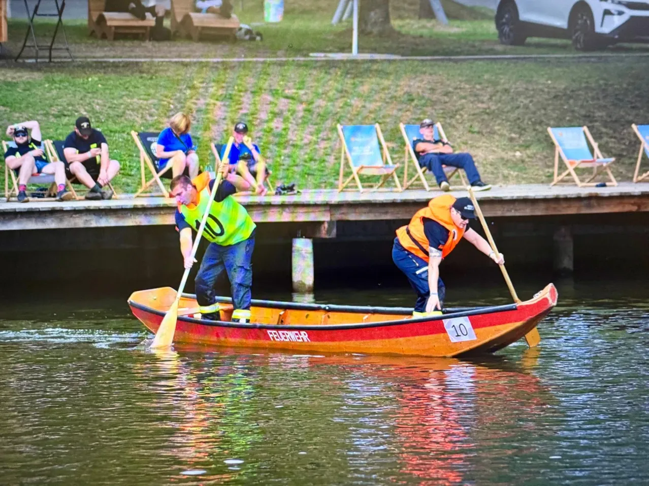Zwei Männer in Warnwesten rudern mit einem roten und orangefarbenen Boot, das mit der Nummer 10 beschriftet ist, auf einem See, während Zuschauer auf Stühlen auf einem Steg im Hintergrund sitzen.