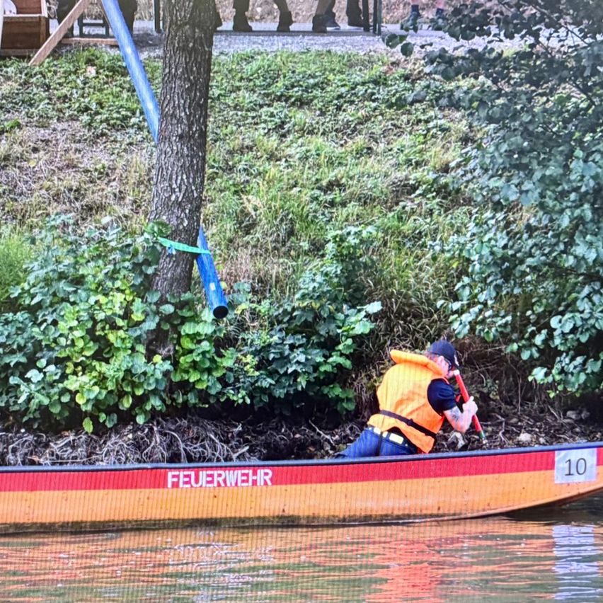 Ein Mann in einem orangefarbenen Rettungsweste paddelt ein Boot mit der Aufschrift 'Feuerwehr' entlang eines Flusses, mit einem Baum und Grünpflanzen im Hintergrund.