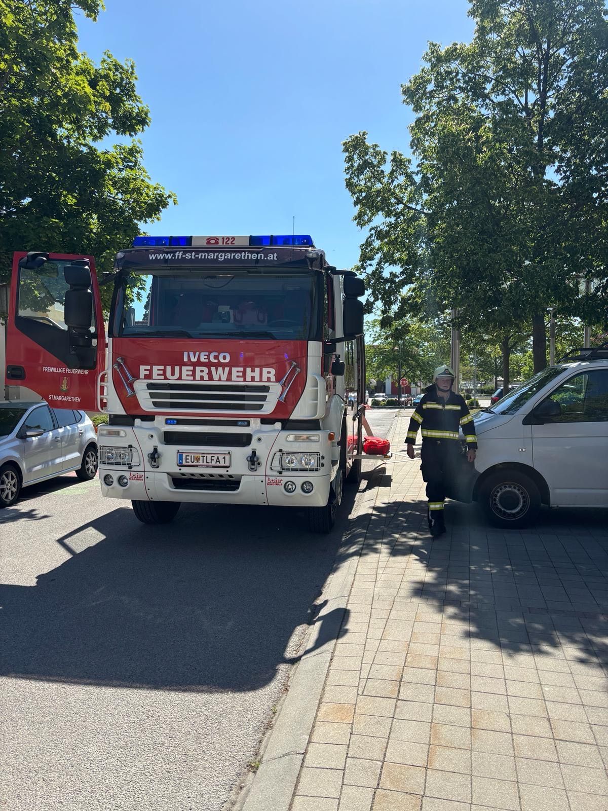 Ein roter Iveco-Feuerwehrwagen mit offenen Türen und ein Feuerwehrmann neben einem weißen Van auf einer Straße.