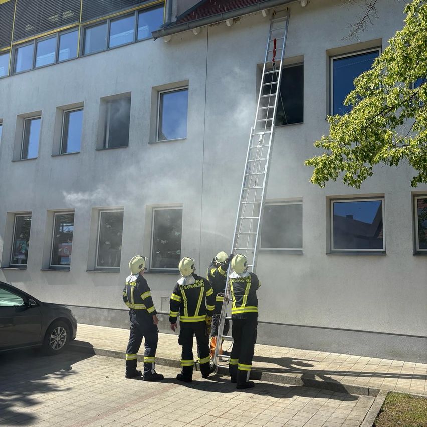 Feuerwehrleute stehen vor einem Gebäude, einer klettert auf eine Leiter. Rauch kommt aus einem Fenster.