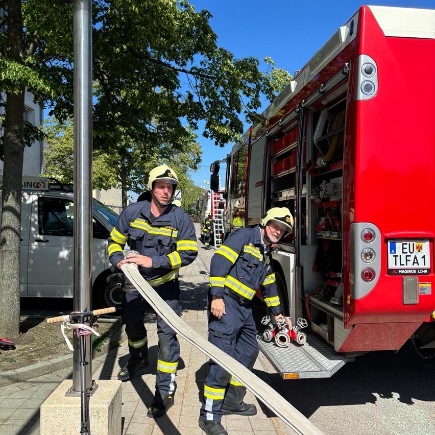 Zwei Feuerwehrleute stehen auf einem Bürgersteig, halten einen Schlauch und bereiten sich darauf vor, ihn an einem Feuerwehrwagen anzubringen. Der Feuerwehrwagen ist rot mit einem Nummernschild, das 'EU TL FA 1' liest. Ein weißer Lieferwagen ist dahinter geparkt.