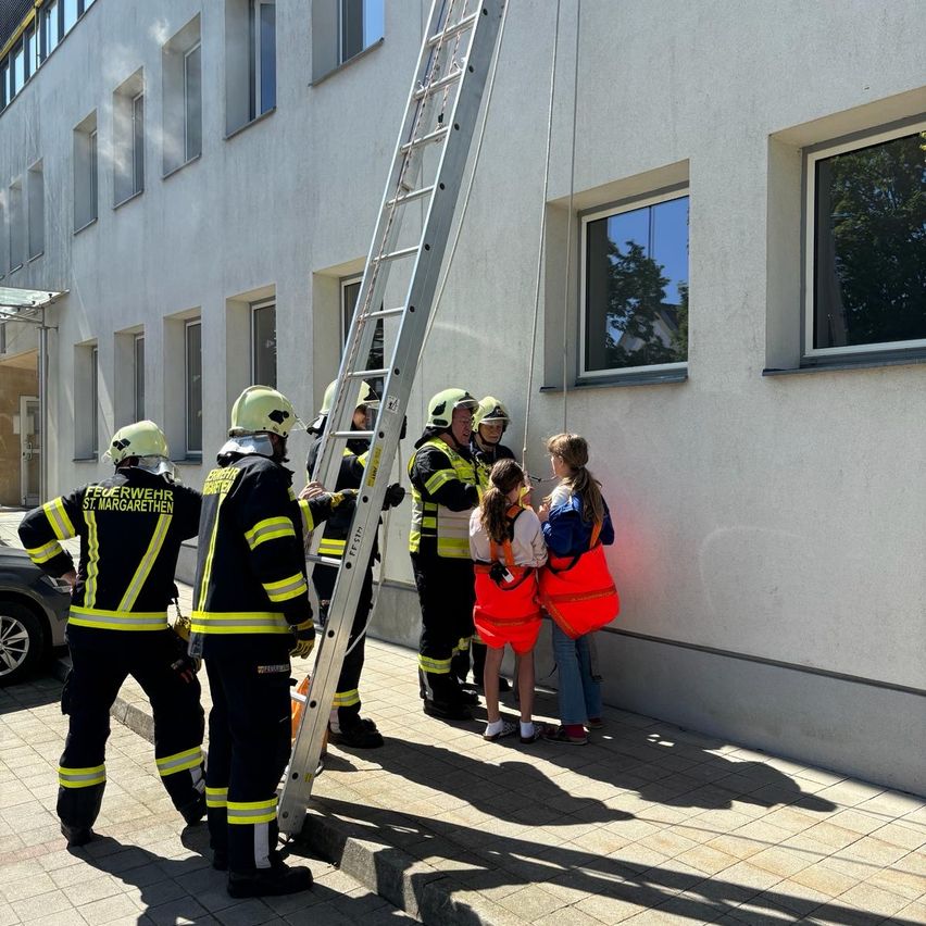 Feuerwehrleute in Uniformen stehen auf einer Leiter, möglicherweise zur Rettung einer Person aus einem Gebäude. In der Nähe stehen eine Frau und ein Mädchen.
