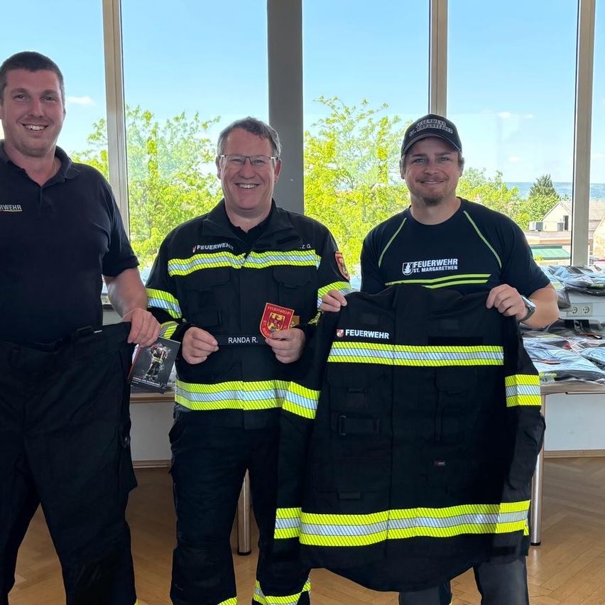 Drei Männer in Feuerwehruniformen posieren für ein Foto und halten eine Feuerwehrjacke hoch. Der Mann in der Mitte hält ein Buch mit einem Bild eines Feuerwehrmanns auf dem Cover. Die Szene spielt sich in einem Raum mit großen Fenstern ab.