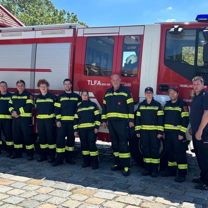 Eine Gruppe von Feuerwehrleuten steht vor einem roten Feuerwehrwagen mit 'TLFA 400-200' an der Seite. Sie tragen alle schwarze Uniformen mit gelben Streifen.