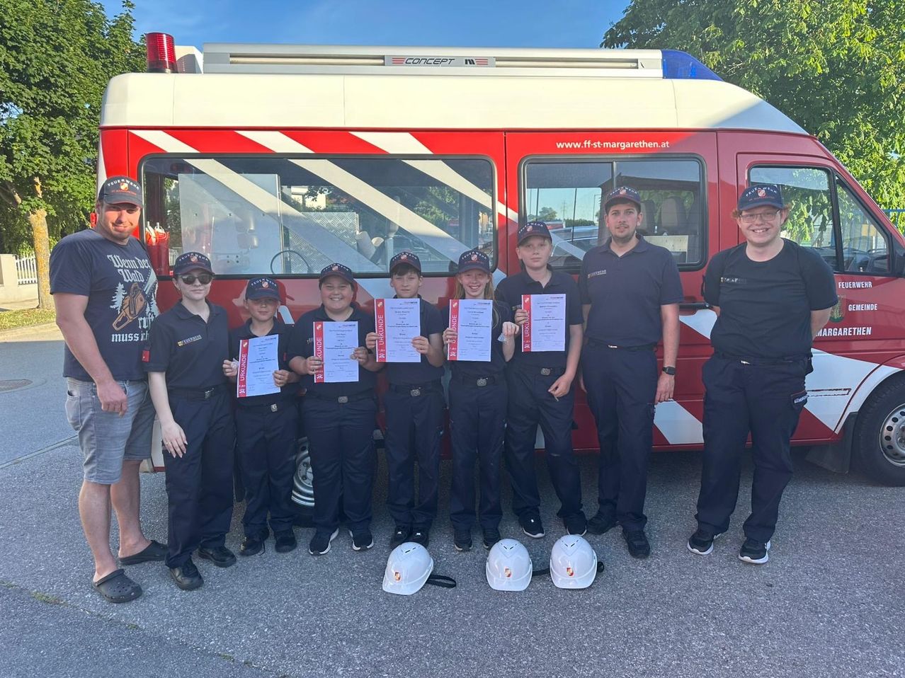 Eine Gruppe von Kindern und Erwachsenen in Feuerwehruniformen posiert mit Zertifikaten vor einem Feuerwehrauto.
