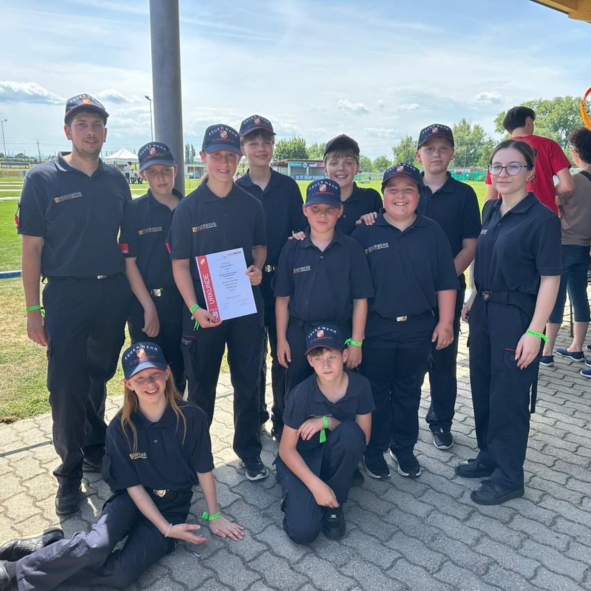 Eine Gruppe von Feuerwehrleuten in Uniform posiert für ein Foto im Freien. Sie lächeln und stehen in einer Reihe, wobei eine Frau ein Zertifikat in der Hand hält.