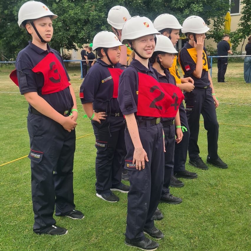 Eine Gruppe von Kindern in Uniformen mit Helmen und nummerierten Westen steht auf einem Grasfeld. Sie scheinen Teil einer organisierten Aktivität oder Veranstaltung zu sein. Bäume und ein Gebäude sind im Hintergrund zu sehen.