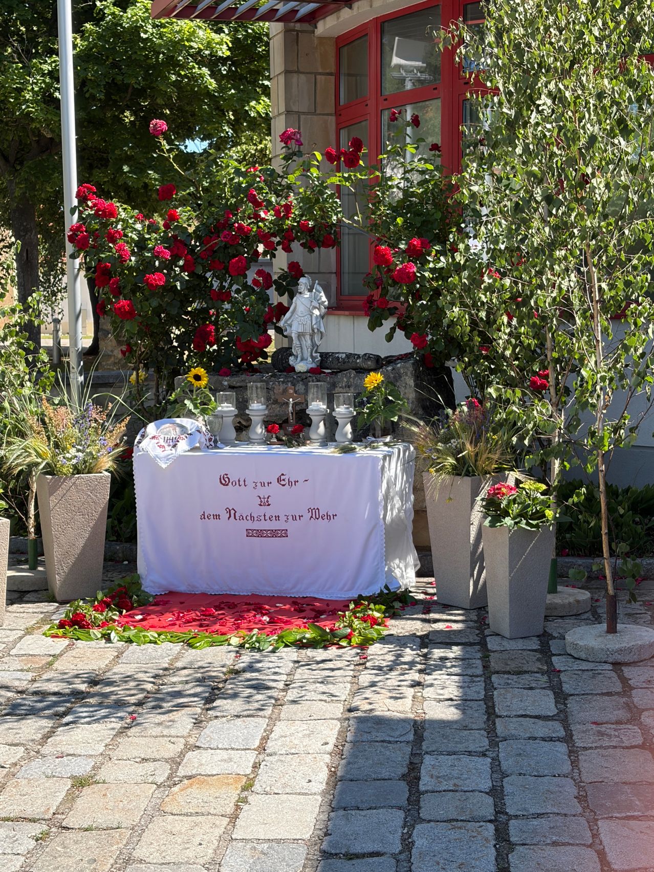 Ein religiöser Altar mit einem weißen Tuch ist mit roten Rosen und Kerzen geschmückt. Er hat eine deutsche Inschrift.