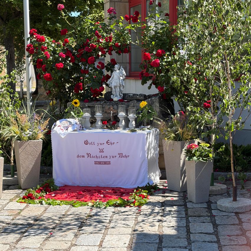 Ein religiöser Altar mit einem weißen Tuch ist mit roten Rosen und Kerzen geschmückt. Er hat eine deutsche Inschrift.
