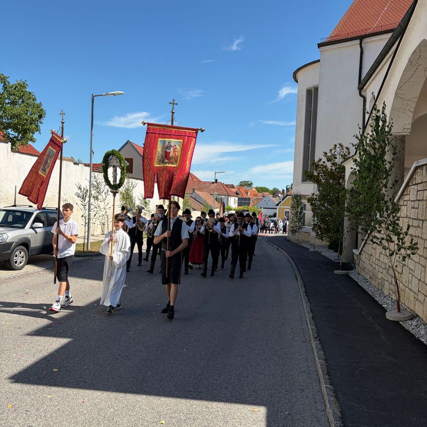 Ein Umzug mit einer Band und Flaggen zieht eine Straße entlang, die von Gebäuden gesäumt ist. Ein Gebäude mit einem roten Dach und einem gewölbten Eingang befindet sich rechts.