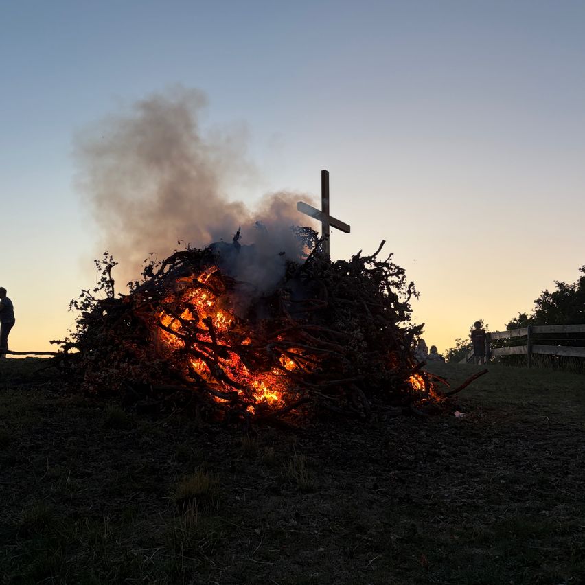 Ein Scheiterhaufen brennt auf einem Hügel, mit einem großen Holzkreuz in der Mitte. Das Feuer ist von trockenen Ästen umgeben und gibt Rauch ab. Menschen stehen in der Nähe.