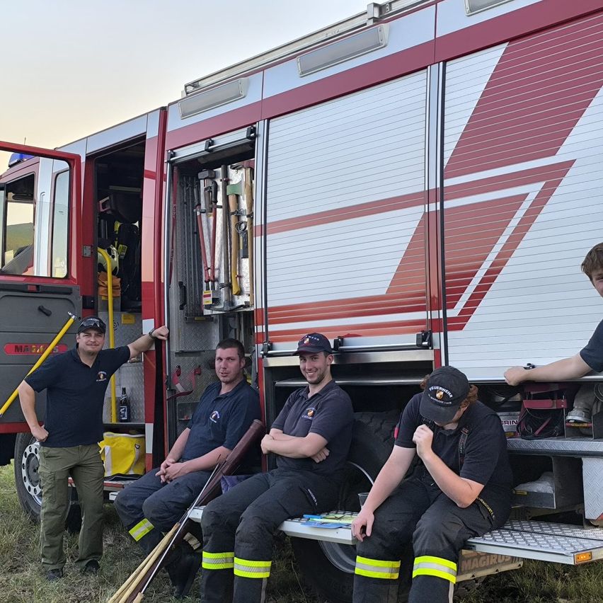 Fünf Feuerwehrleute sitzen und stehen neben einem Feuerwehrauto. Zwei sitzen auf der Stufe des Trucks, einer mit einer Hand am Kinn, während die anderen beiden auf dem Boden sitzen und nach oben schauen. Der fünfte Mann steht neben der offenen Tür und hält einen Stock.