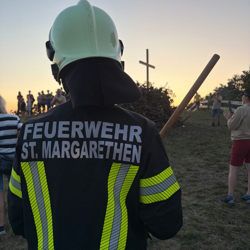 Ein Feuerwehrmann aus St. Margarethen steht mit einer Menschenmenge, die ein Lagerfeuer beobachtet. Der Himmel ist orange, während die Sonne untergeht.