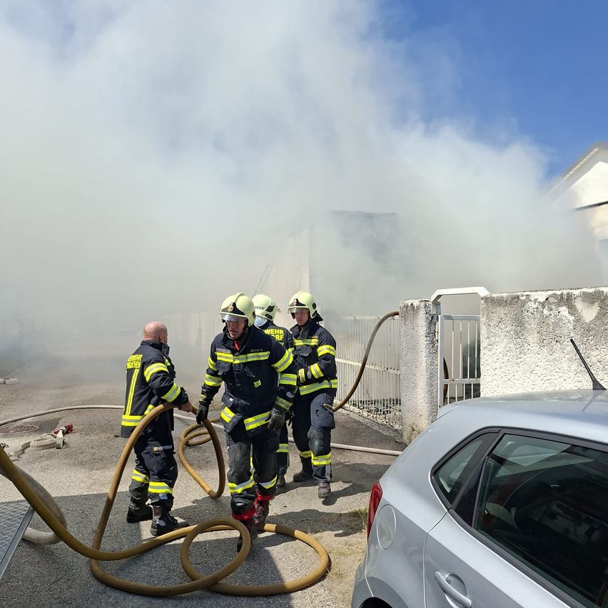 Feuerwehrleute bekämpfen einen Gebäudebrand, wobei Rauch aus dem Dach aufsteigt. Einer hält einen Schlauch, während zwei andere beobachten. Ein silbernes Auto ist in der Nähe geparkt.