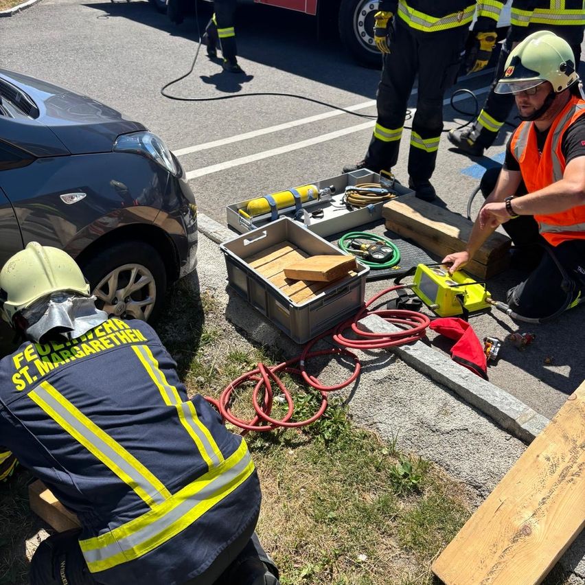 Feuerwehrleute arbeiten an einem Auto, einer kniet neben einer Werkzeugkiste und Werkzeugen auf dem Boden. Ein weiterer Feuerwehrmann ist in der Nähe, trägt eine Uhr und eine reflektierende Weste.
