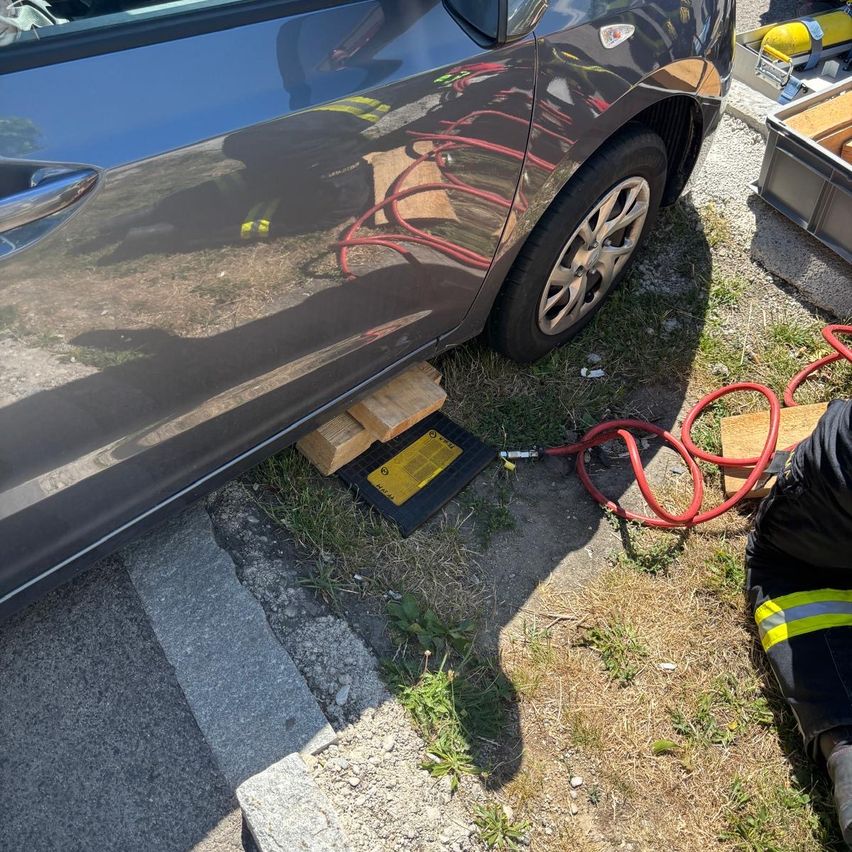 Ein Feuerwehrmann arbeitet an einem Auto, das mit einem Wagenheber unter ihm steht. Das Auto ist auf Gras geparkt, und ein roter Schlauch ist in der Nähe.