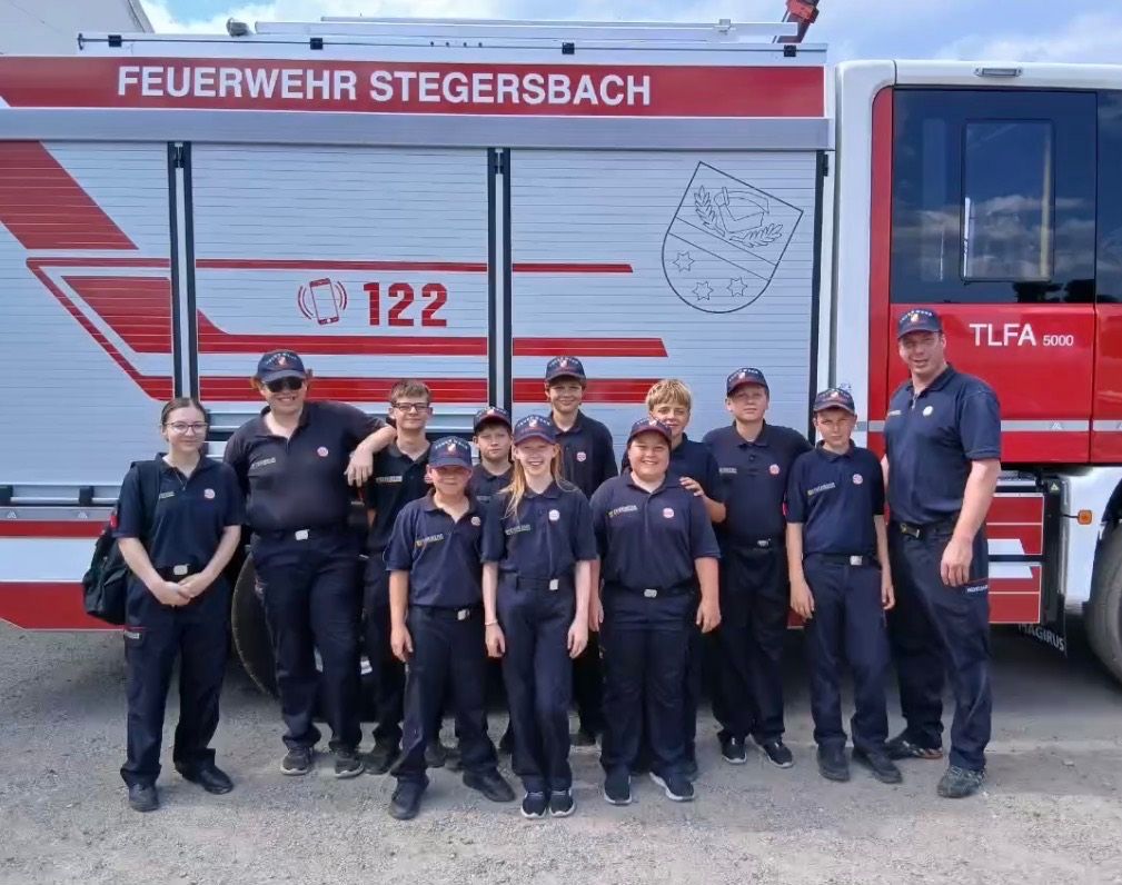 Eine Gruppe Feuerwehrleute in Uniform, einschließlich Kinder, steht vor einem Feuerwehrauto mit der Aufschrift 'Feuerwehr Stegersbach 122.' Sie alle lächeln für ein Gruppenfoto.