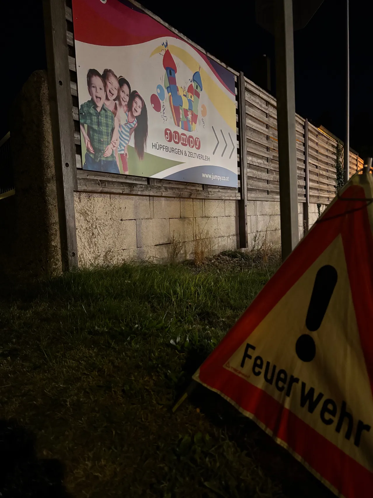 Eine große Werbung für eine Kinderveranstaltung ist an einem Holzzaun angebracht. Die Werbung zeigt eine Gruppe von Kindern, die lächeln, und enthält den Text 'Jump' und 'Hupburgen & Zeltverleih'.