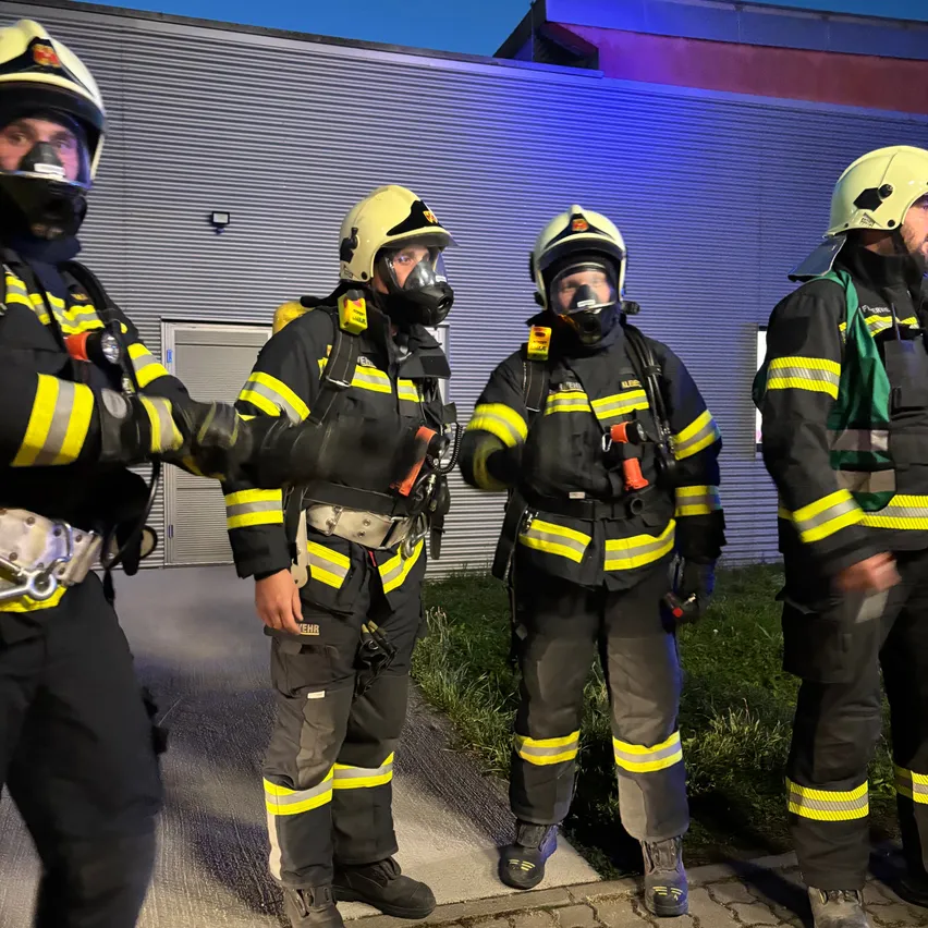 Fünf Feuerwehrleute in voller Ausrüstung, einschließlich Helmen und Masken, stehen nachts vor einem Gebäude.