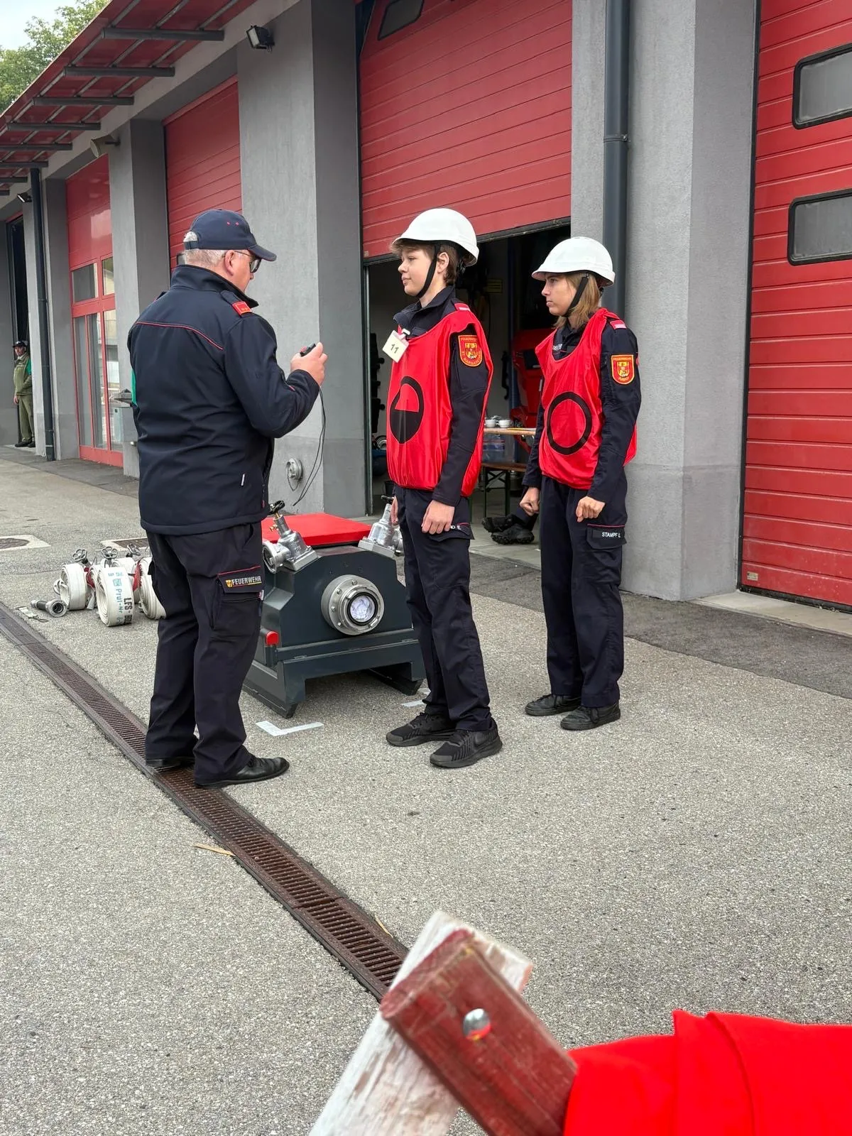 Drei Personen in roten und schwarzen Uniformen stehen vor einer Feuerwache.