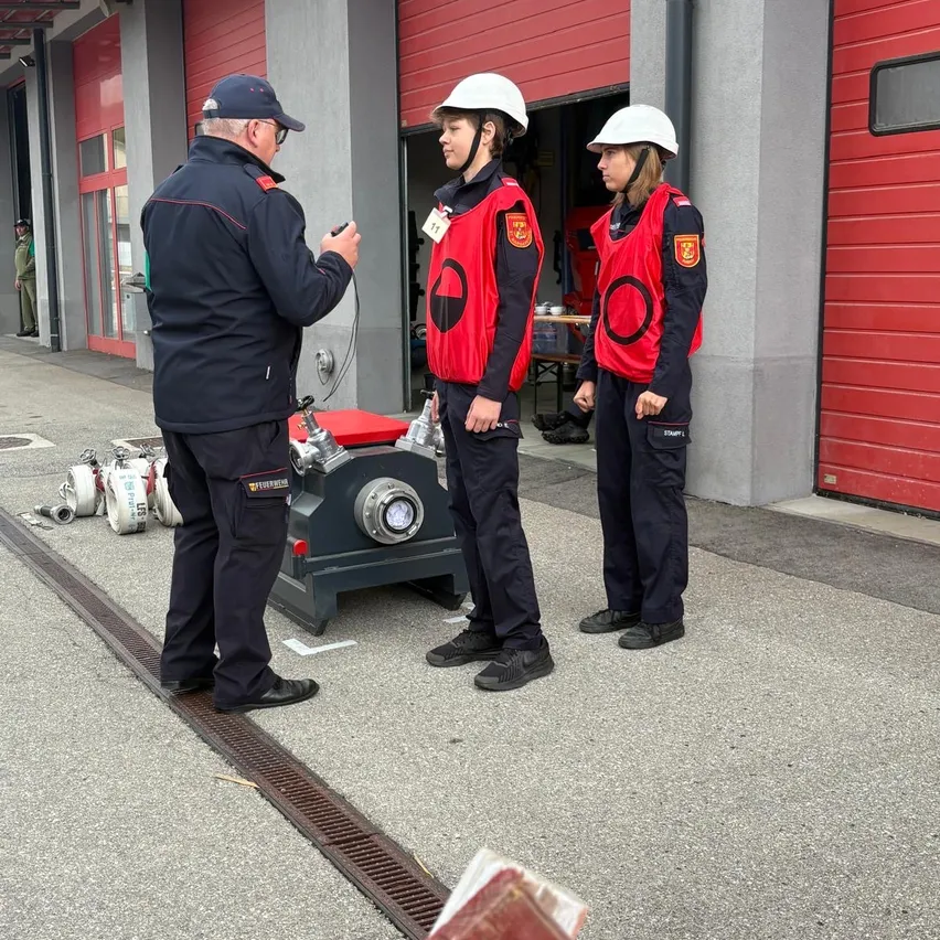 Drei Personen in roten und schwarzen Uniformen stehen vor einer Feuerwache.