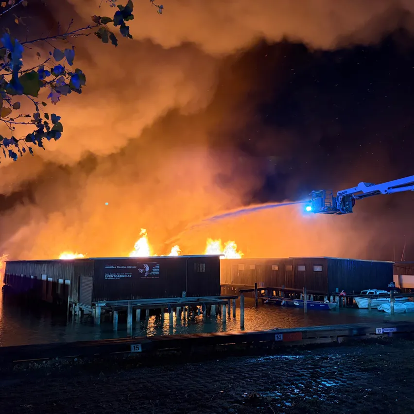 Ein Gebäude auf einem Dock brennt nachts. Ein Feuerwehrwagen spritzt Wasser auf das Feuer.