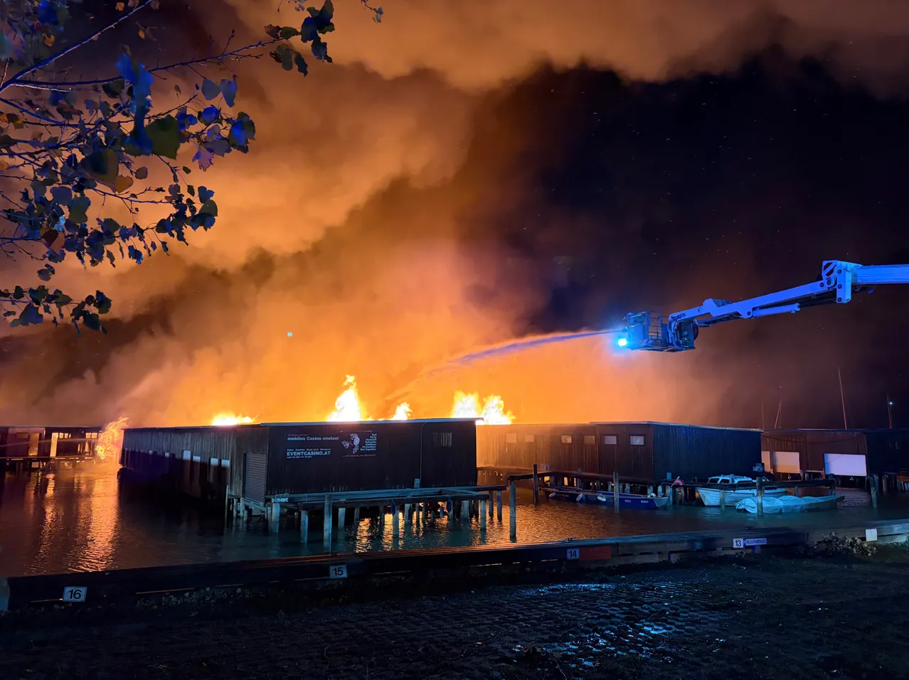 Ein Gebäude auf einem Dock brennt nachts. Ein Feuerwehrwagen spritzt Wasser auf das Feuer.