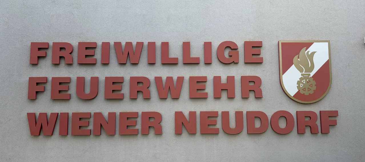 Ein Schild an einer Wand mit der Aufschrift 'Freiwillige Feuerwehr Wiener Neudorf' und einem Logo mit einer Flamme und einem Zahnrad.