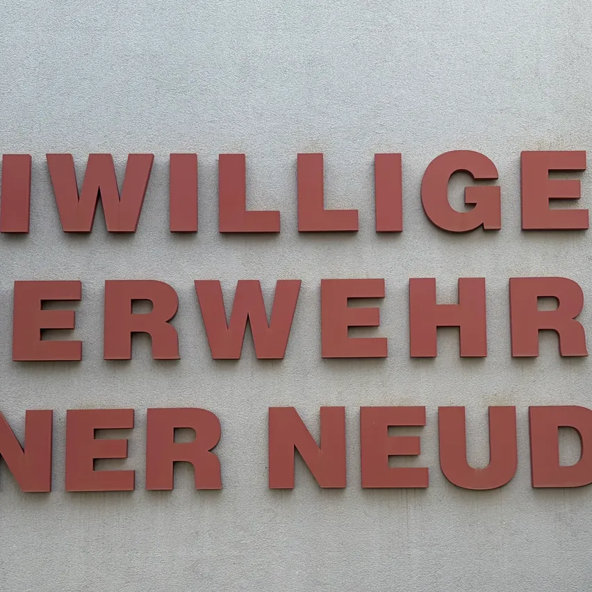 Ein Schild an einer Wand mit der Aufschrift 'Freiwillige Feuerwehr Wiener Neudorf' und einem Logo mit einer Flamme und einem Zahnrad.