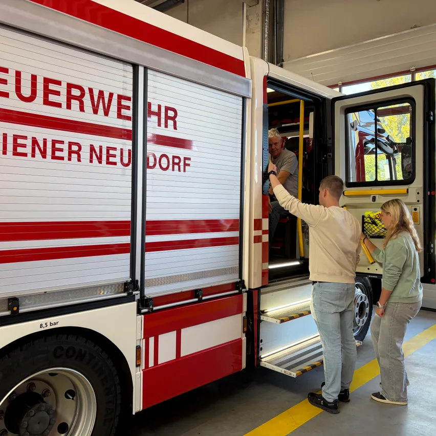 Ein Mann und eine Frau stehen neben einem Feuerwehrauto mit offener Tür. Der Mann zeigt auf das Auto, während die Frau ein gelbes Objekt hält.