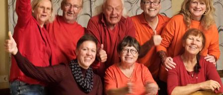 Eine Gruppe von Erwachsenen in passenden roten und orangefarbenen Shirts posiert für ein Foto mit Lächeln und Daumen hoch.
