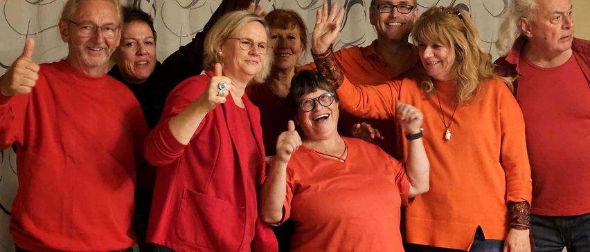 A group of adults in orange shirts and jeans pose for a photo, giving thumbs up. The room has white curtains and a chandelier.
