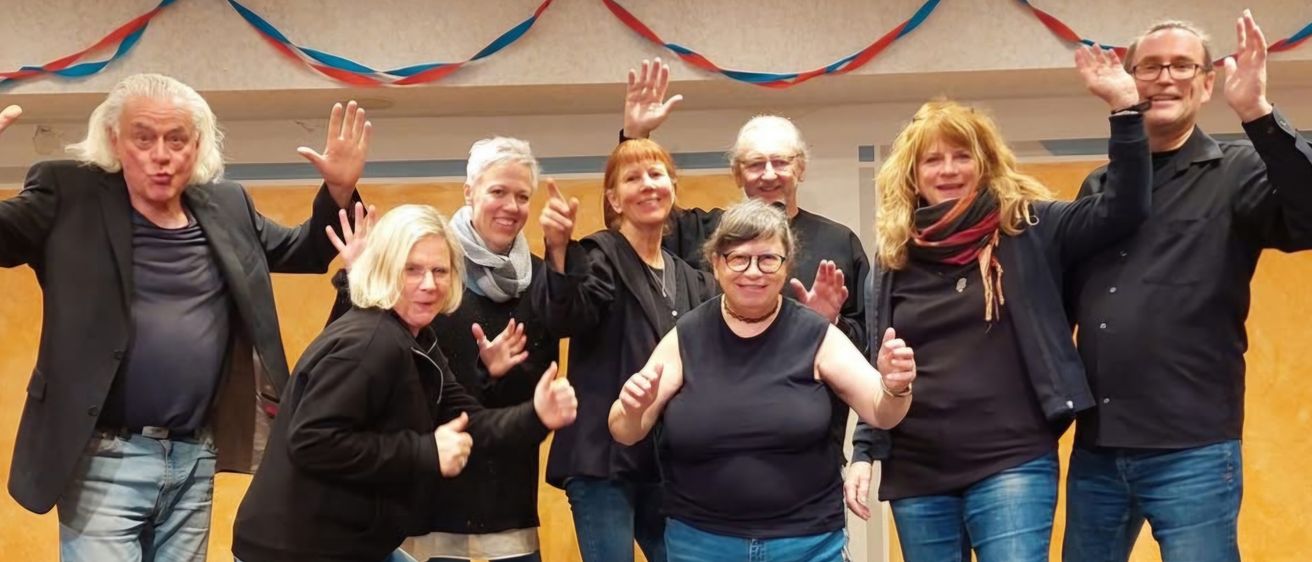 Eine Gruppe von Erwachsenen, sowohl Männer als auch Frauen, posiert für ein Foto in einem Raum mit gelber Wand und dekorativem Wimpelband. Sie tragen alle Jacken, Jeans und Turnschuhe. Einige von ihnen machen Handgesten.