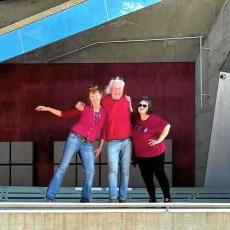 Drei Menschen posieren auf einer Terrasse für ein Foto, wobei eine Frau ihre Arme ausstreckt. Hinter ihnen ist eine rote Wand.