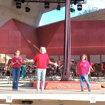 Drei Personen auf der Bühne mit roten Shirts, vor einem Orchester stehend, bei roten Lichtern auftretend.