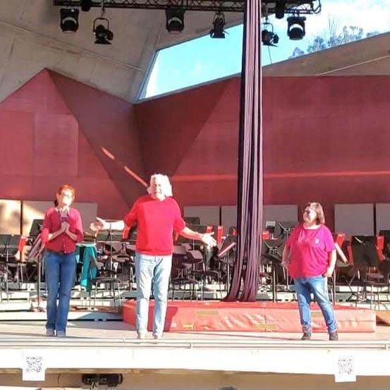 Drei Personen auf der Bühne mit roten Shirts, vor einem Orchester stehend, bei roten Lichtern auftretend.