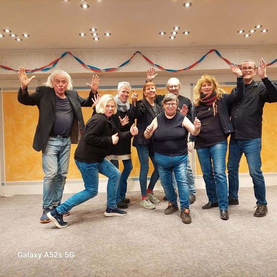 Eine Gruppe von Menschen in Schwarz posiert für ein Foto mit erhobenen Händen in einem Raum mit gelber Wand und bunten Dekorationen.
