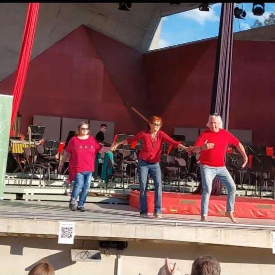 Drei Erwachsene, zwei Frauen und ein Mann, treten auf der Bühne auf, mit einem Orchester im Hintergrund. Die Bühne ist vor einer roten Kulisse und hat eine rote Polsterung.