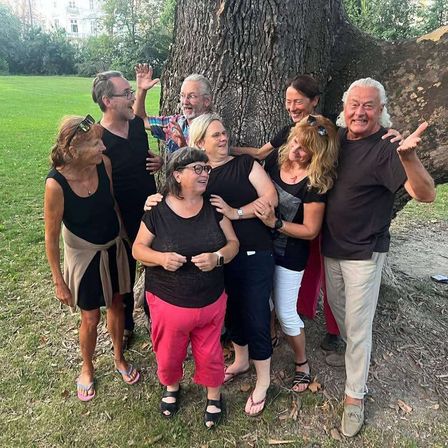 Eine Gruppe von Erwachsenen steht im Kreis, lächelt und posiert für ein Foto. Sie stehen vor einem großen Baum in einem Park.