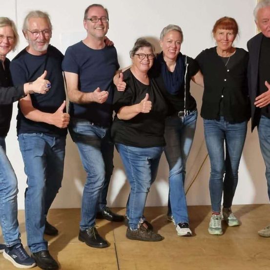 Eine Gruppe von Menschen steht eng beieinander für ein Foto, alle lächeln. Sie sind in Freizeitkleidung gekleidet, einschließlich Jeans und Shirts, und einige tragen Brillen. Der Hintergrund ist eine einfache weiße Wand.