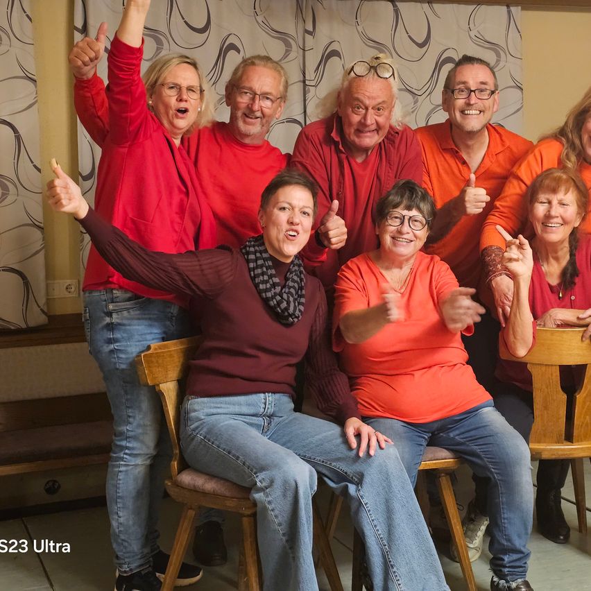 Eine Gruppe von Menschen in passenden orangefarbenen und roten Oberteilen, sitzend und stehend, posiert für ein Foto. Sie alle lächeln und machen Handzeichen. Der Hintergrund zeigt einen gemusterten Vorhang.