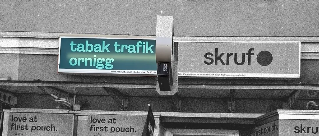 Ein Schwarz-Weiß-Foto einer Ladenfront mit einem Schild, das 'tabak trafik ornigg' und ein weiteres Schild, das 'skruf' liest. Es gibt mehrere Maschinen und Poster an der Vorderseite des Ladens.
