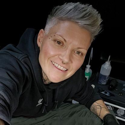 Eine Frau mit grauem Haar lächelt für ein Selfie, trägt eine schwarze Kapuzenjacke und grüne Shorts. Sie hat einen Nasenring und Tattoos an den Armen. Im Hintergrund sind Flaschen und ein Plastikbeutel zu sehen.